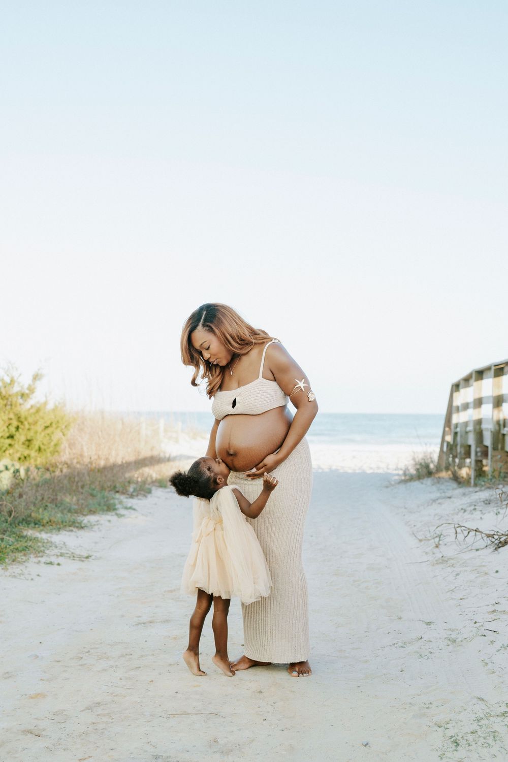 Pregnant woman in cream dress with young daughter kissing her baby bump on a sandy beach path.