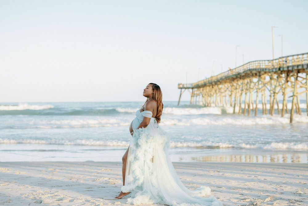 Pregnant woman in flowing white gown poses on sunlit beach near pier with ocean waves behind her.