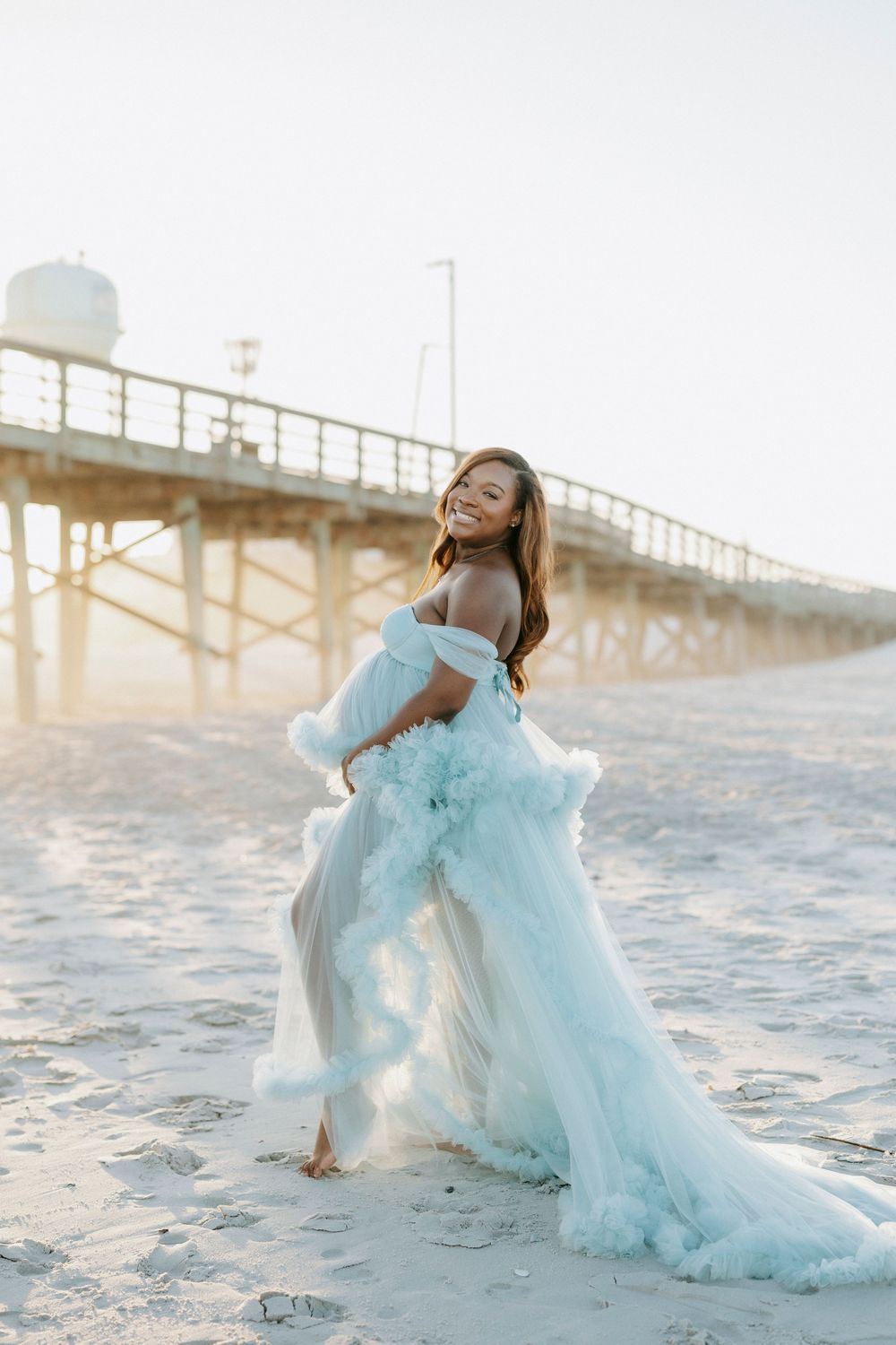 Smiling pregnant woman in flowing light blue tulle gown poses on sandy beach near wooden pier at sunset.