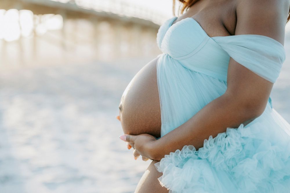Pregnant woman in light blue ruffled dress holding baby bump on beach at sunset.