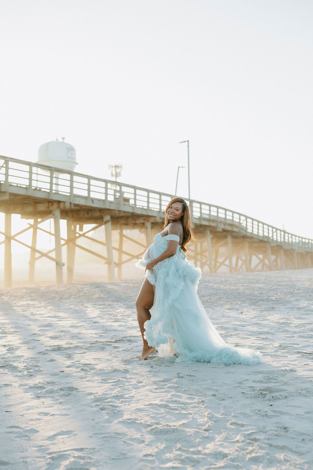 Pregnant woman in elegant light blue off-shoulder tulle maternity gown stands gracefully on beach by pier.