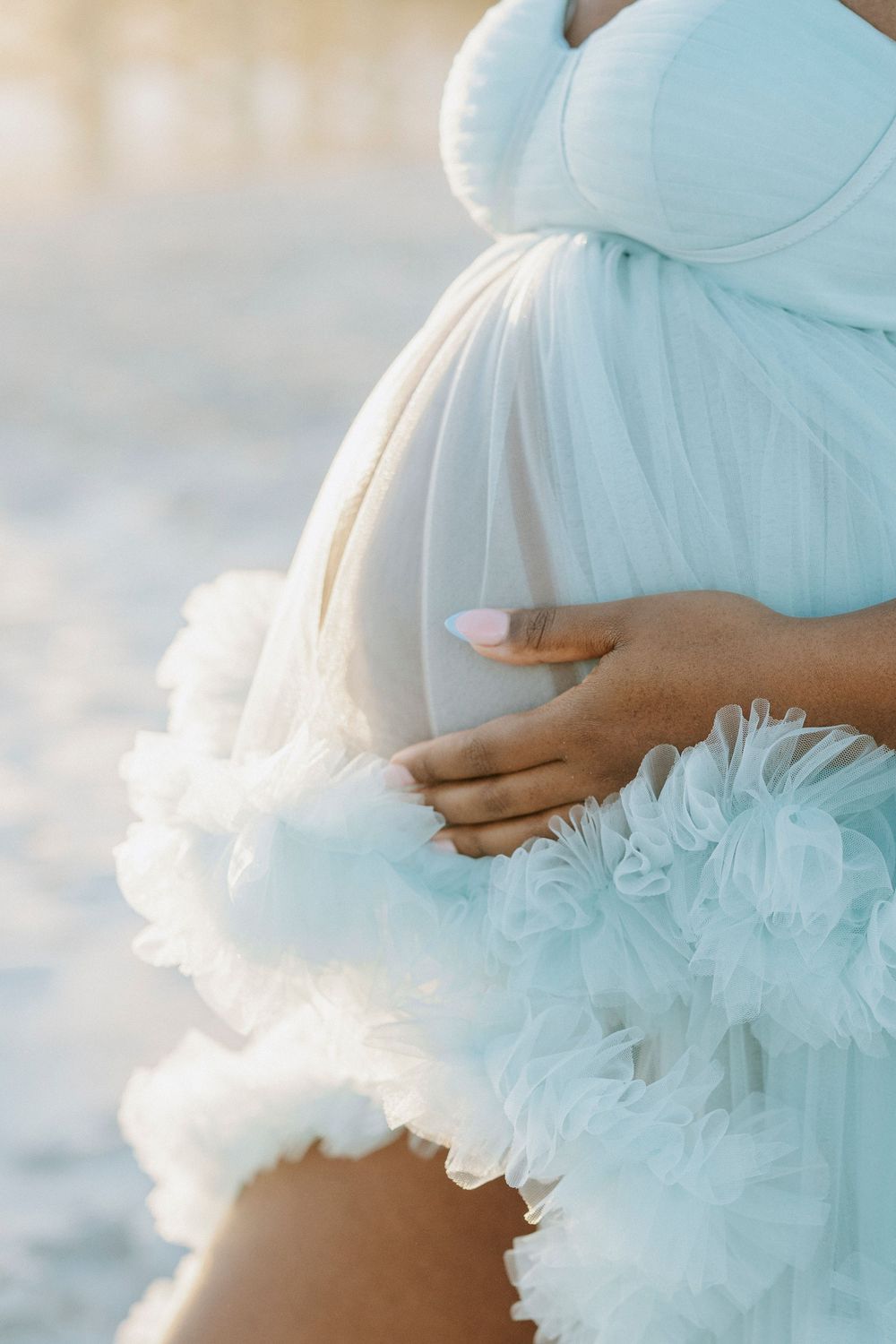 Pregnant woman in light blue ruffled gown cradling baby bump with pink manicured nails at golden hour.