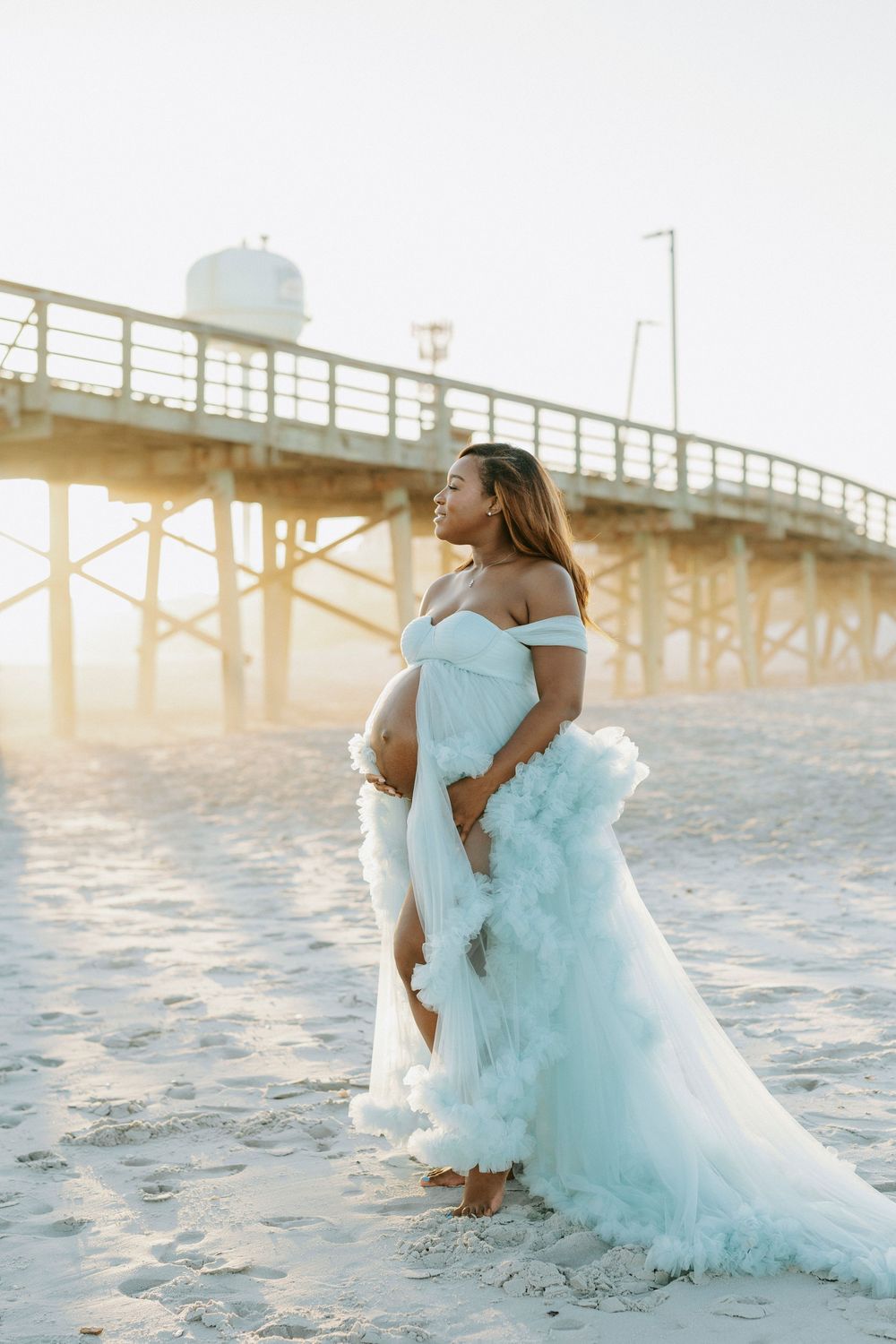 Expectant mother in white ruffled tulle gown cradles baby bump on sunlit beach near wooden pier.