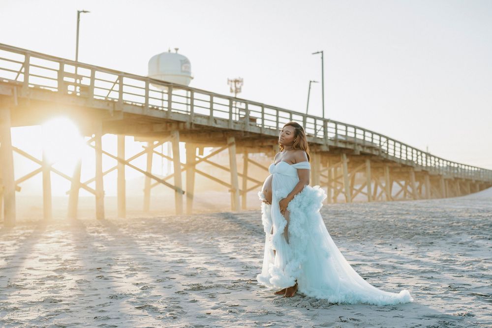 Bride in white gown standing on beach near pier at golden sunset, dress train flowing in soft light.