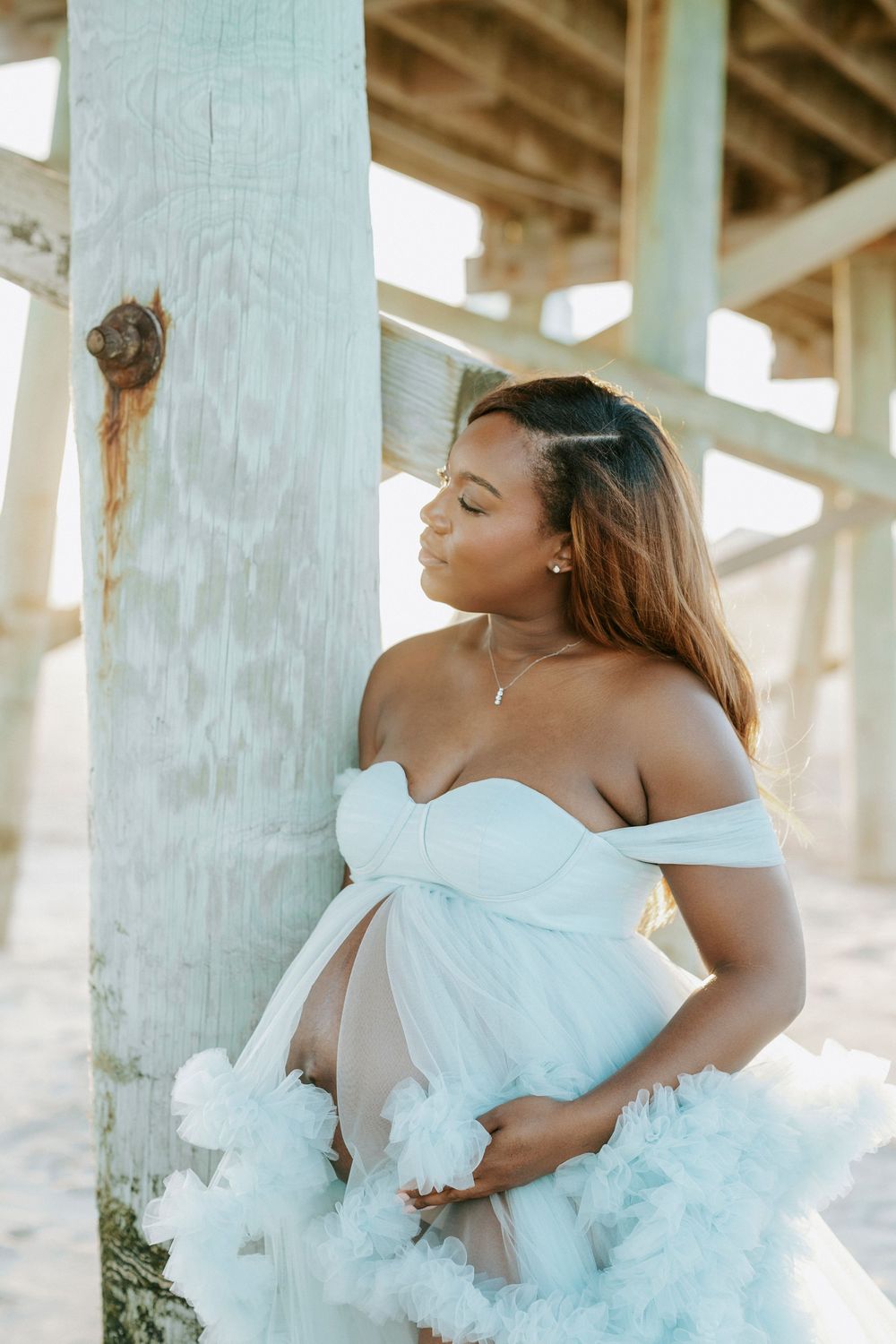 Pregnant woman in white off-shoulder tulle gown poses under beach pier, cradling baby bump in soft natural light.