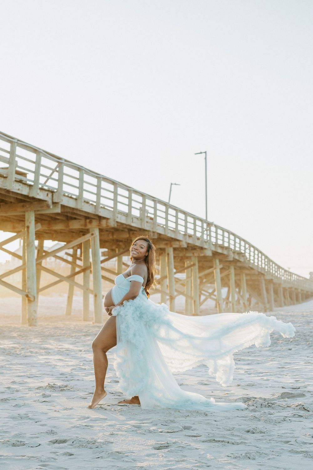 Pregnant woman in flowing light blue tulle gown poses on sandy beach near wooden pier at sunset.