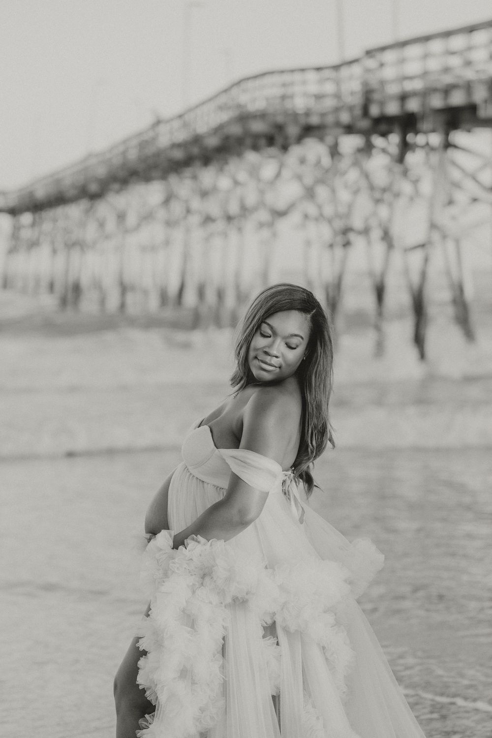 Black and white photo of pregnant woman in flowing tulle gown posing near ocean pier on beach.