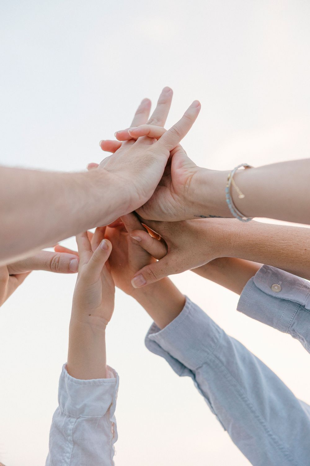 Group of people stacking hands together outdoors, symbolizing teamwork, unity, and collaboration against a bright sky.