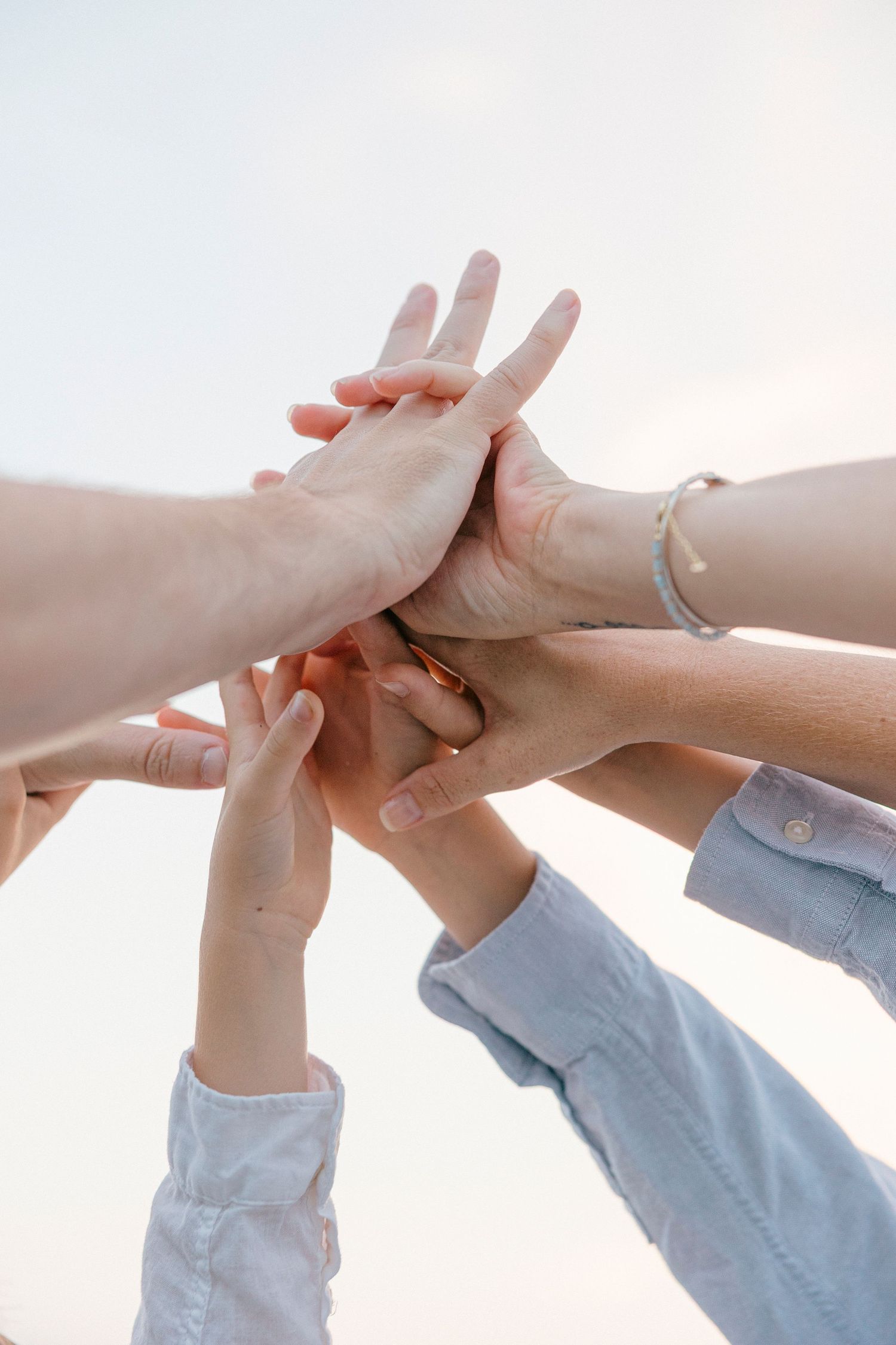 Group of people stacking hands together outdoors, symbolizing teamwork, unity, and collaboration against a bright sky.
