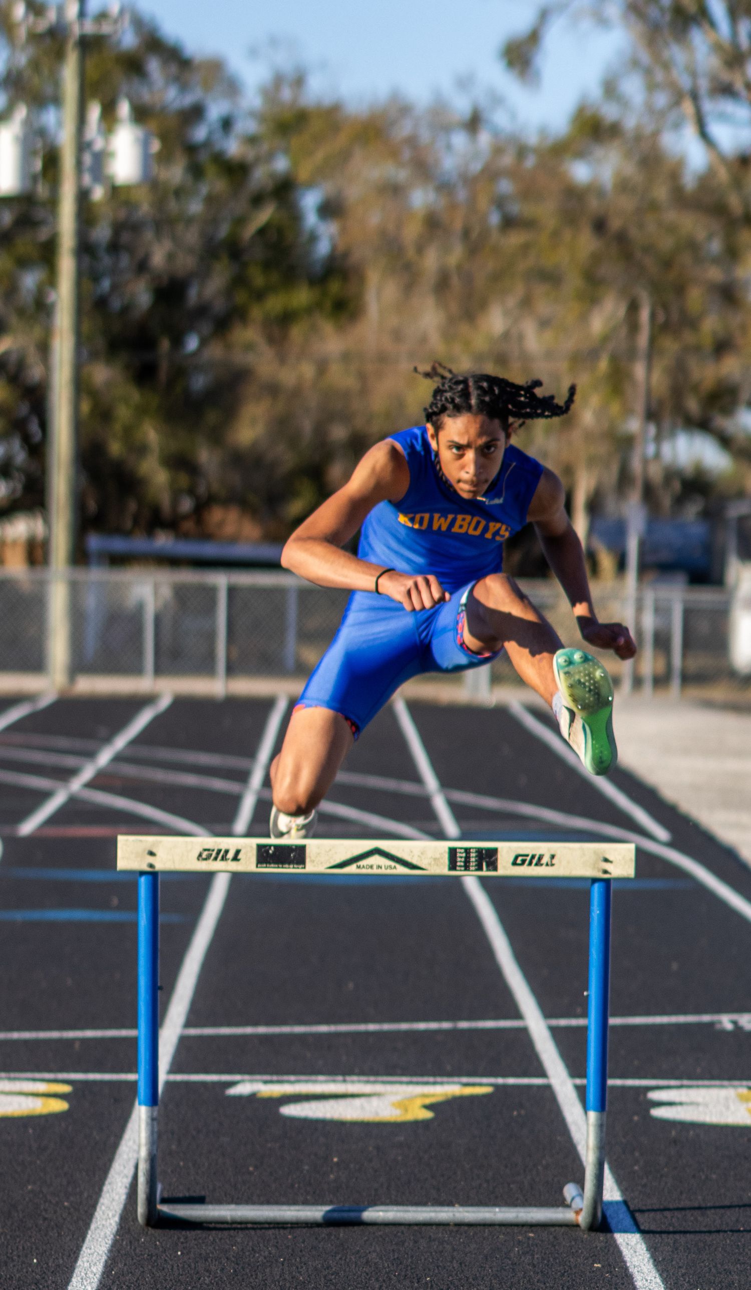 Track athlete in blue uniform clears hurdles during practice session on outdoor running track.