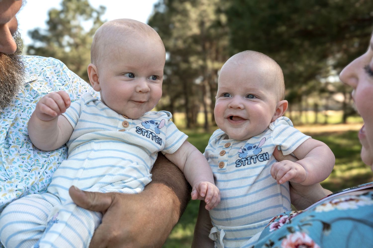 A baby wearing a striped onesie smiles while sitting outdoors in a grassy area on a sunny day.