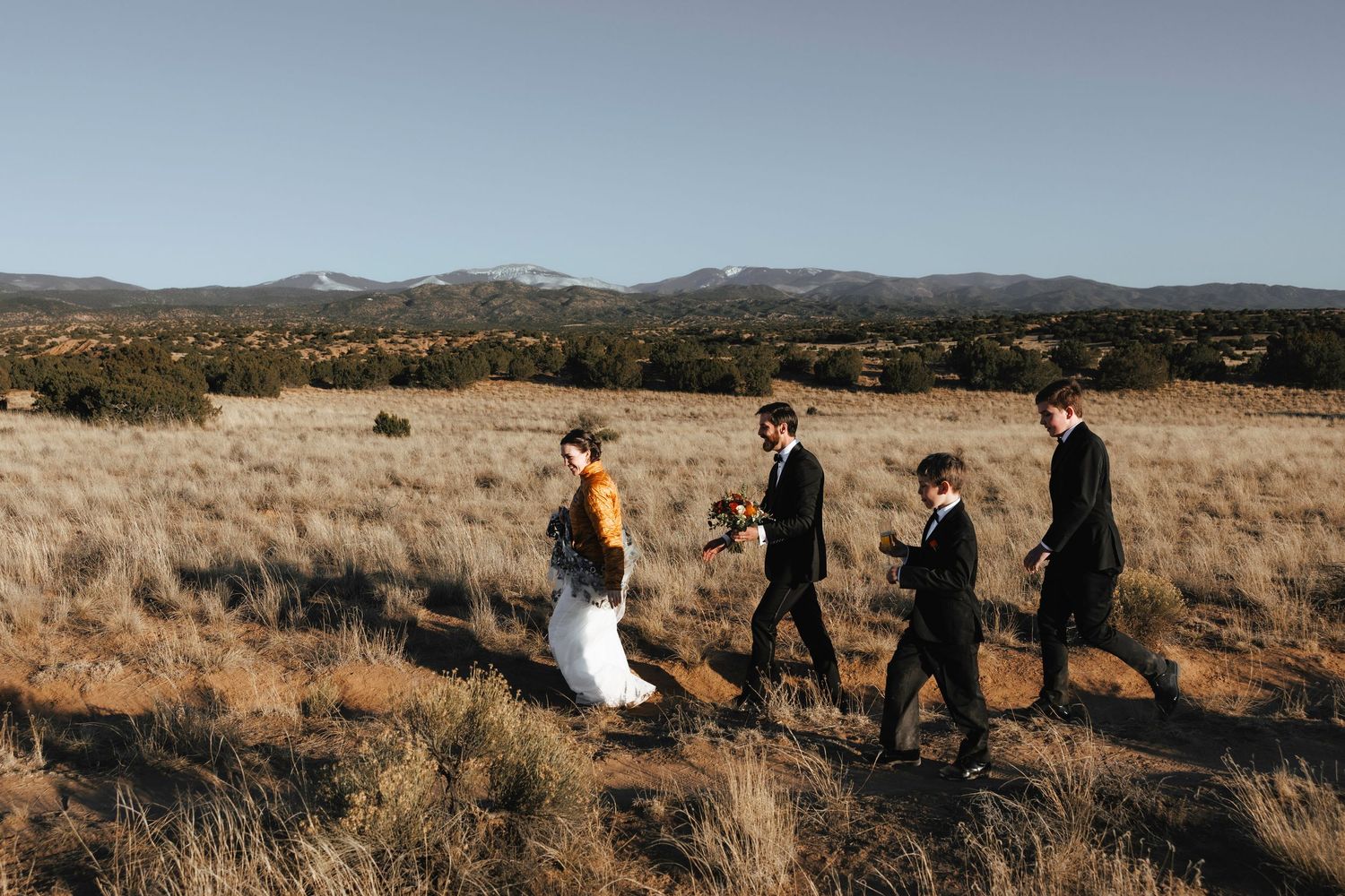 Bride and groom walking with wedding party through desert landscape with mountain views.