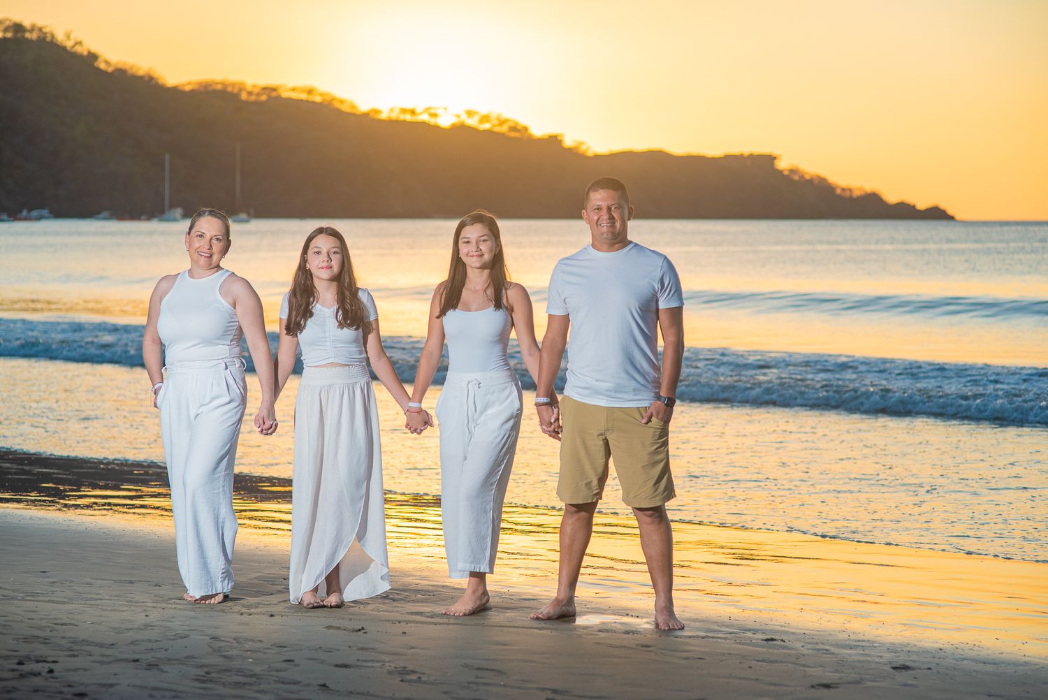 Family of four holding hands on a sandy beach at golden sunset, wearing coordinating white and blue outfits.