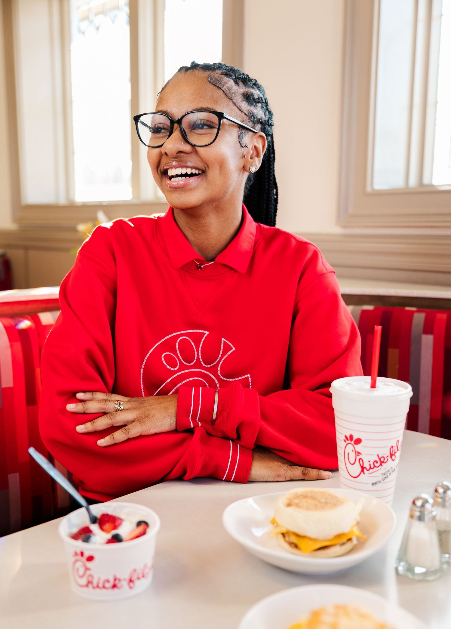 A person in a red sweatshirt sits at a table with a drink and plate of food, laughing and smiling warmly.