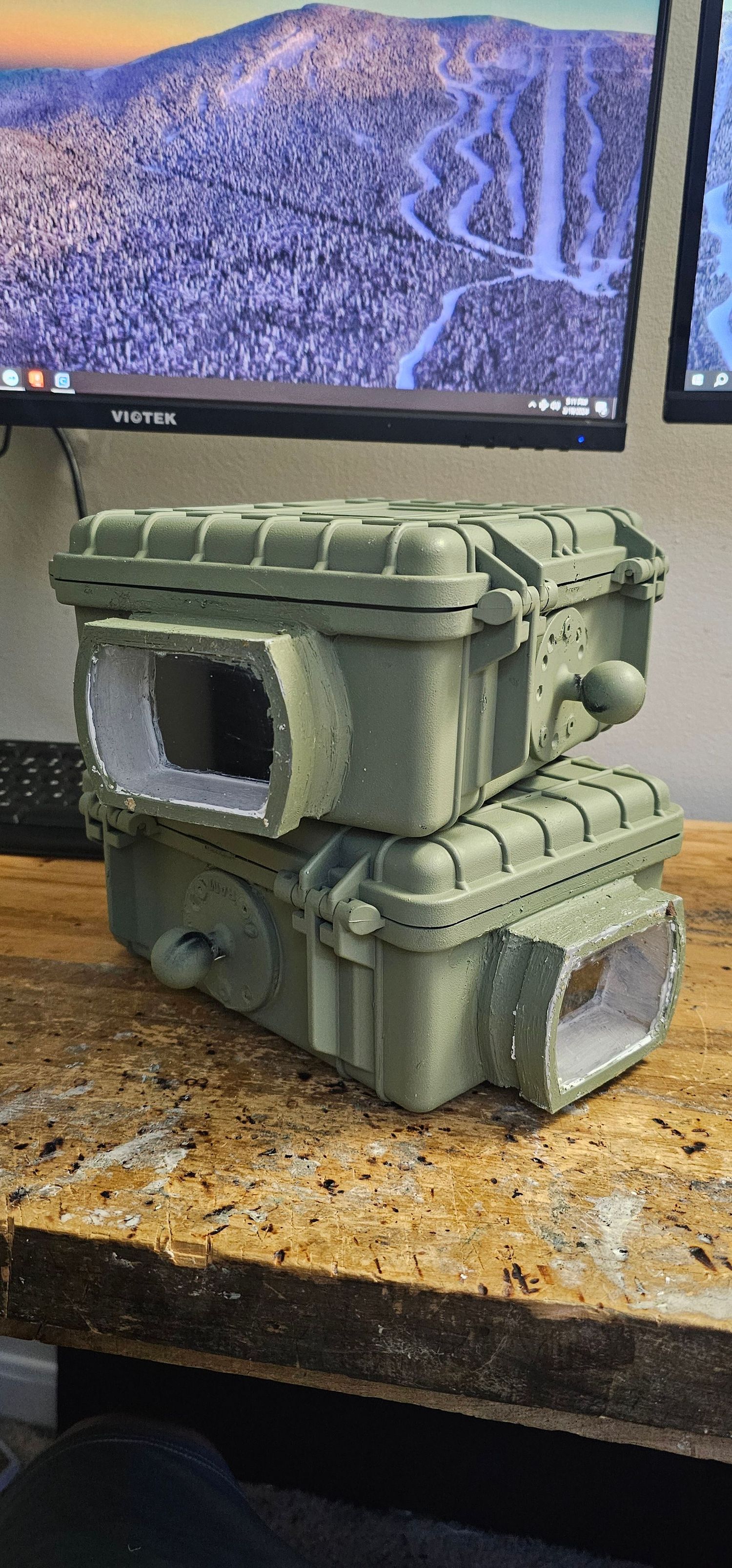 Two olive green Pelican case DIY wildlife camera traps stacked on a workbench indoors.