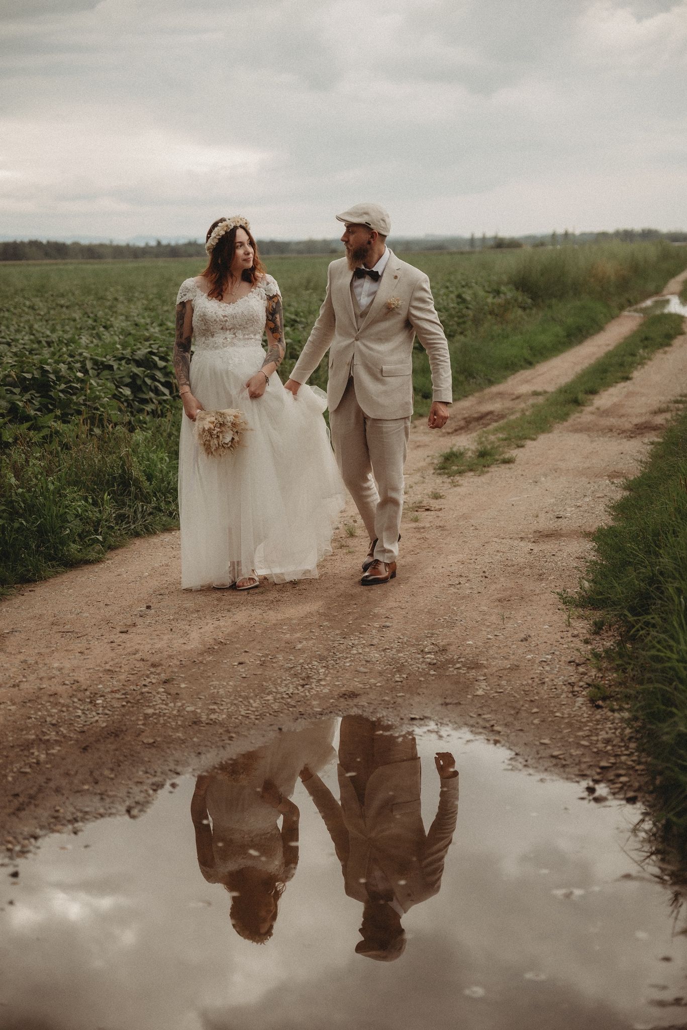 Bride and groom walking a dirt road reflected in a puddle on their wedding day.