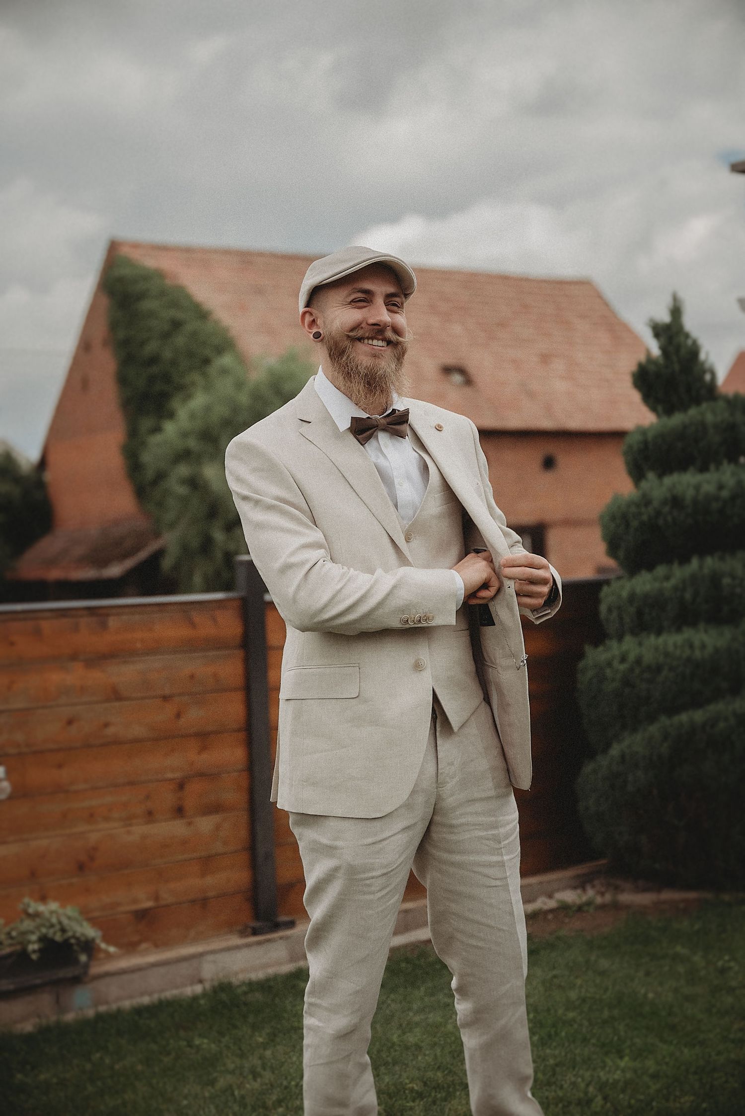Bearded groom in cream linen suit and flat cap smiling outdoors at a wedding venue.