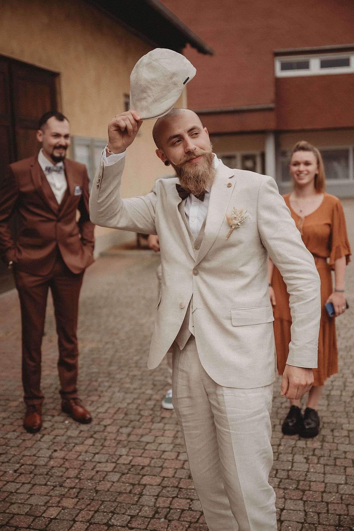 Stylish groom in cream suit tipping his flat cap outdoors at a wedding celebration.