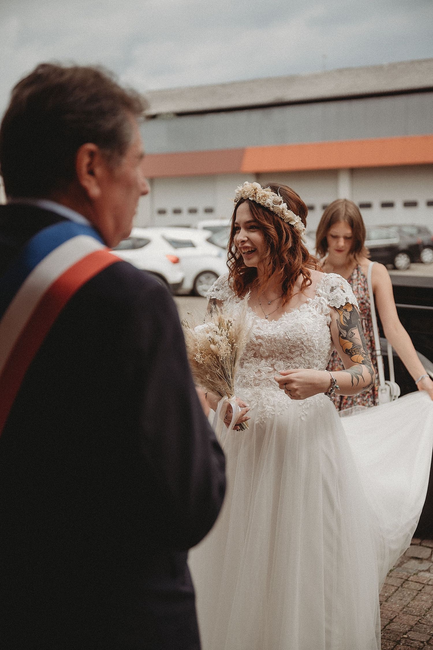 Smiling bride in floral crown arrives at civil ceremony greeted by official with tricolor sash.
