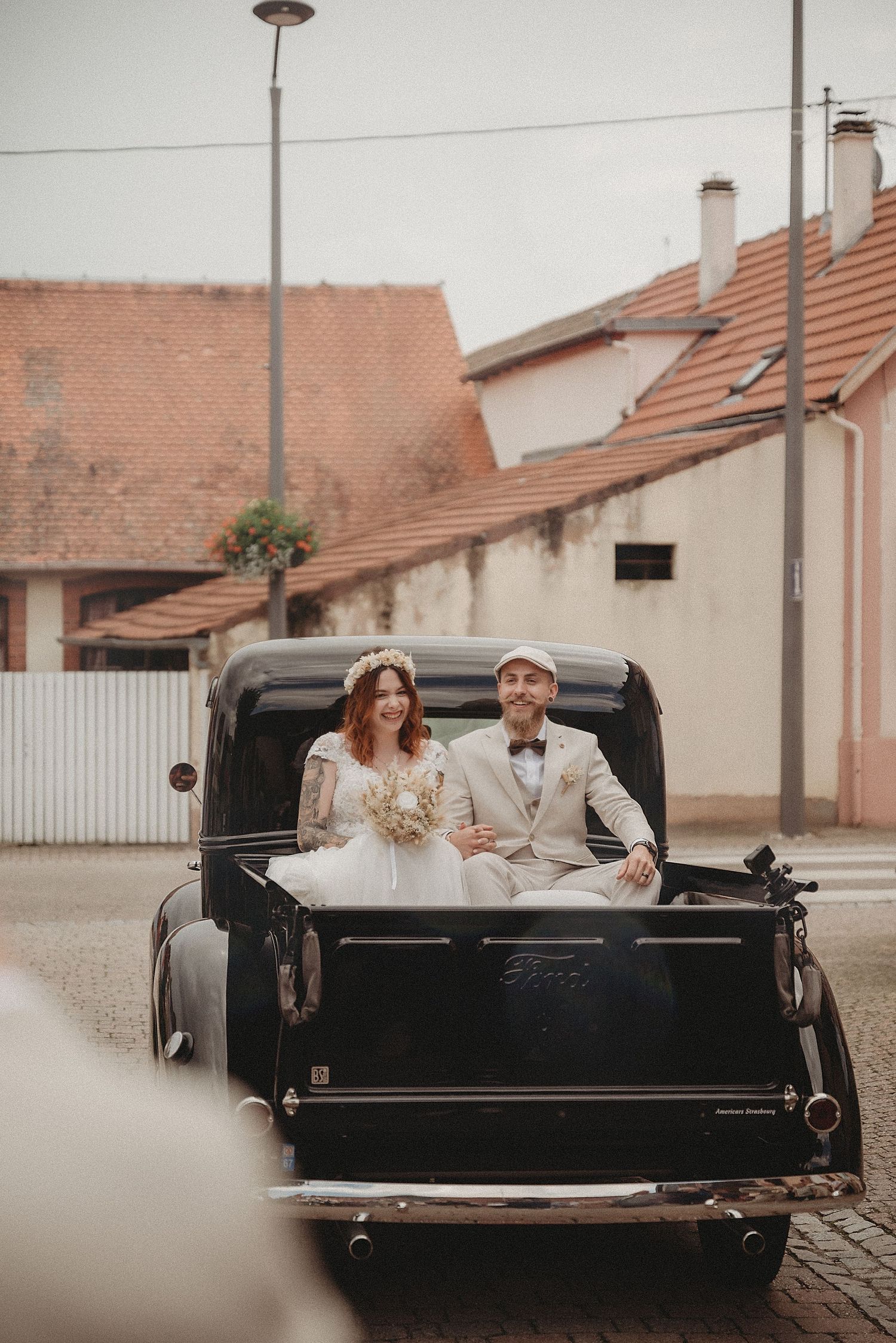 Newlyweds ride in the back of a vintage black Ford truck through a charming European village street.