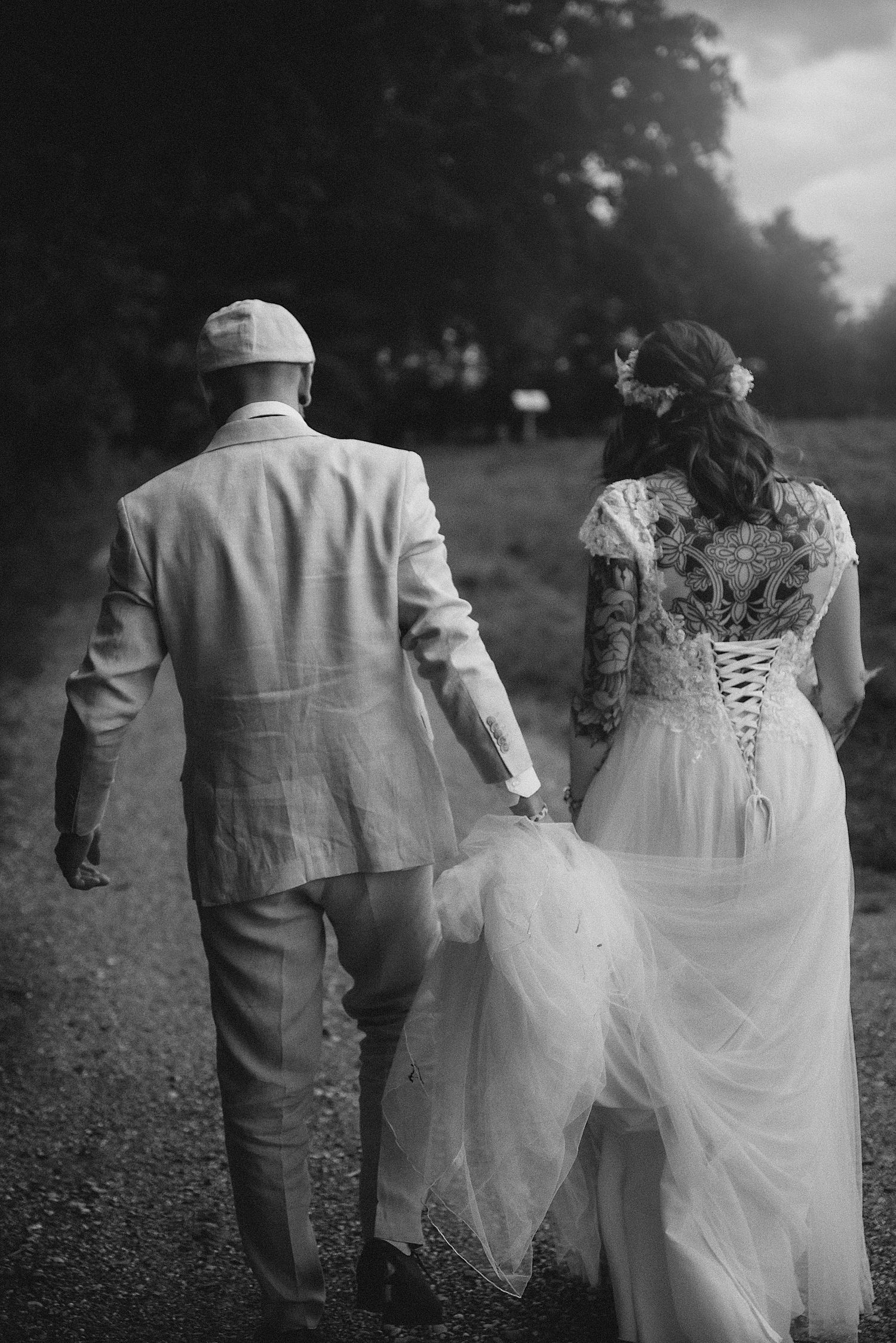 Black and white photo of bride and groom walking hand in hand, bride in lace corset wedding gown viewed from behind.