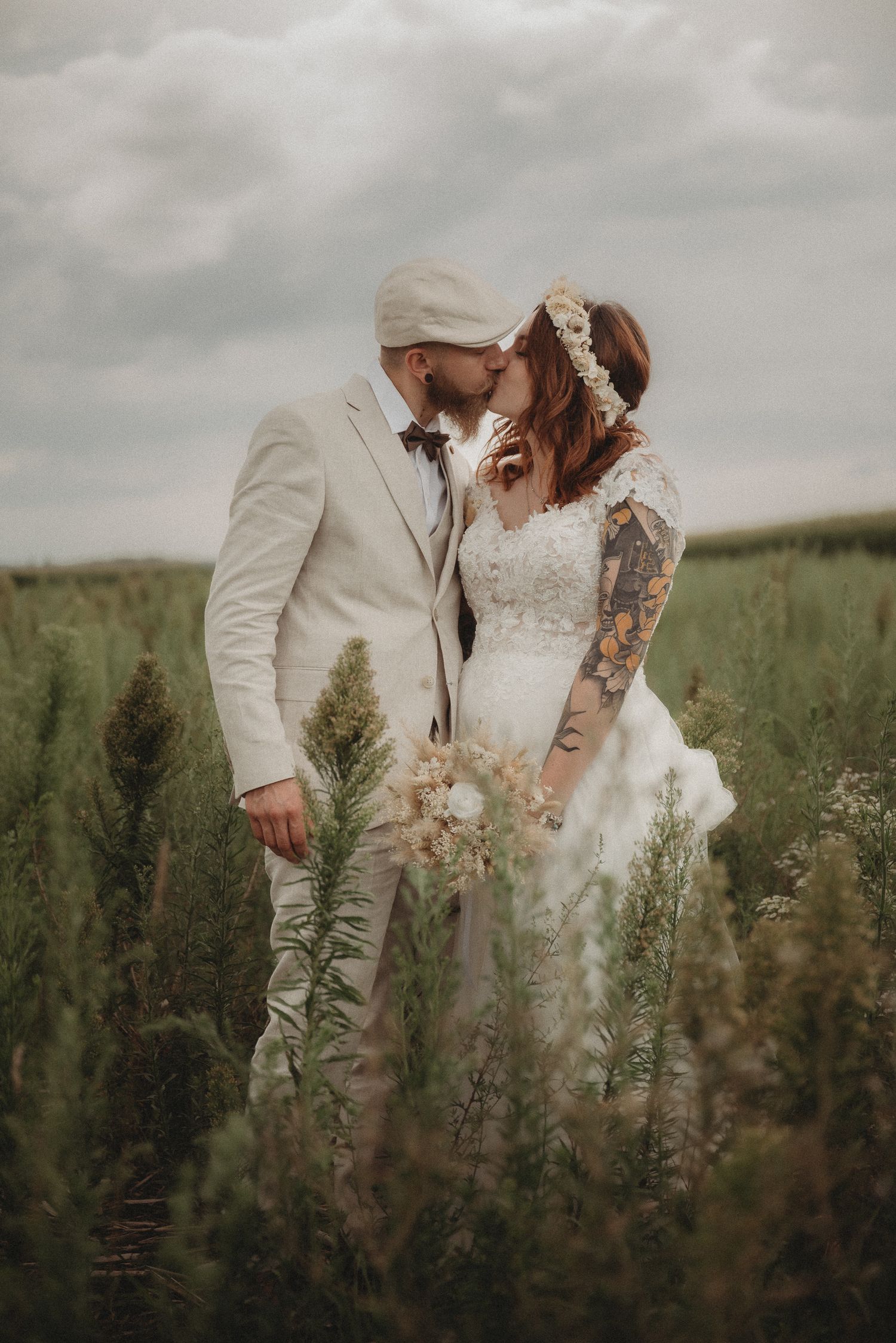 Bride and groom sharing a romantic kiss in a green meadow, bride in lace gown with floral crown and bouquet.