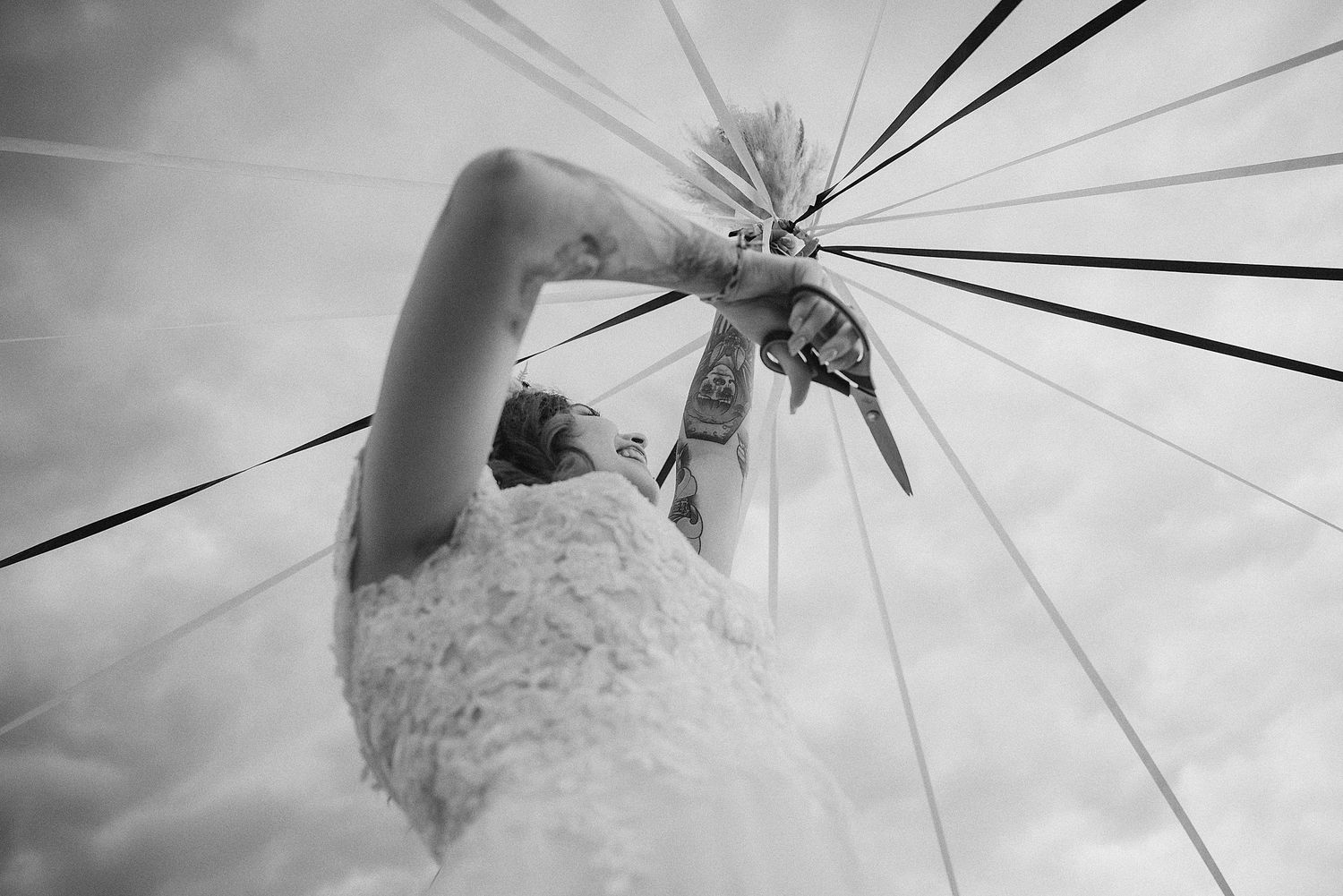 Black and white low angle shot of bride holding ribbon bouquet against dramatic cloudy sky.