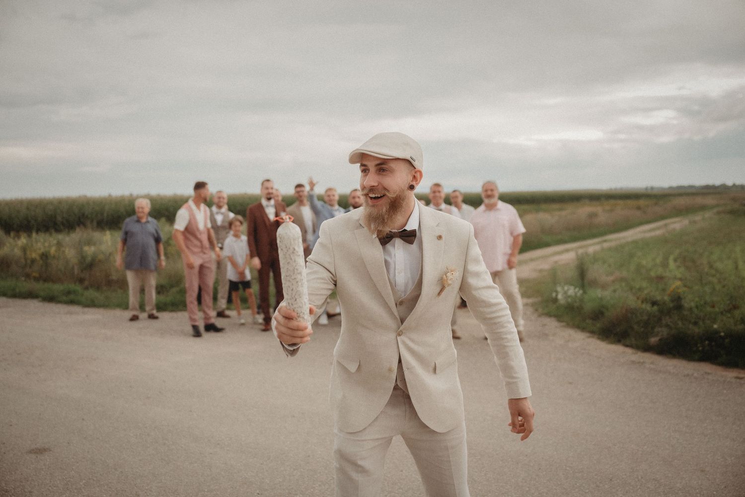 Groom in beige suit and flat cap laughs joyfully with groomsmen gathered on a rural road.