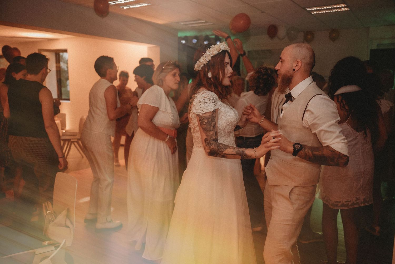 Bride in floral crown dances with tattooed groom and guests at colorful wedding reception.