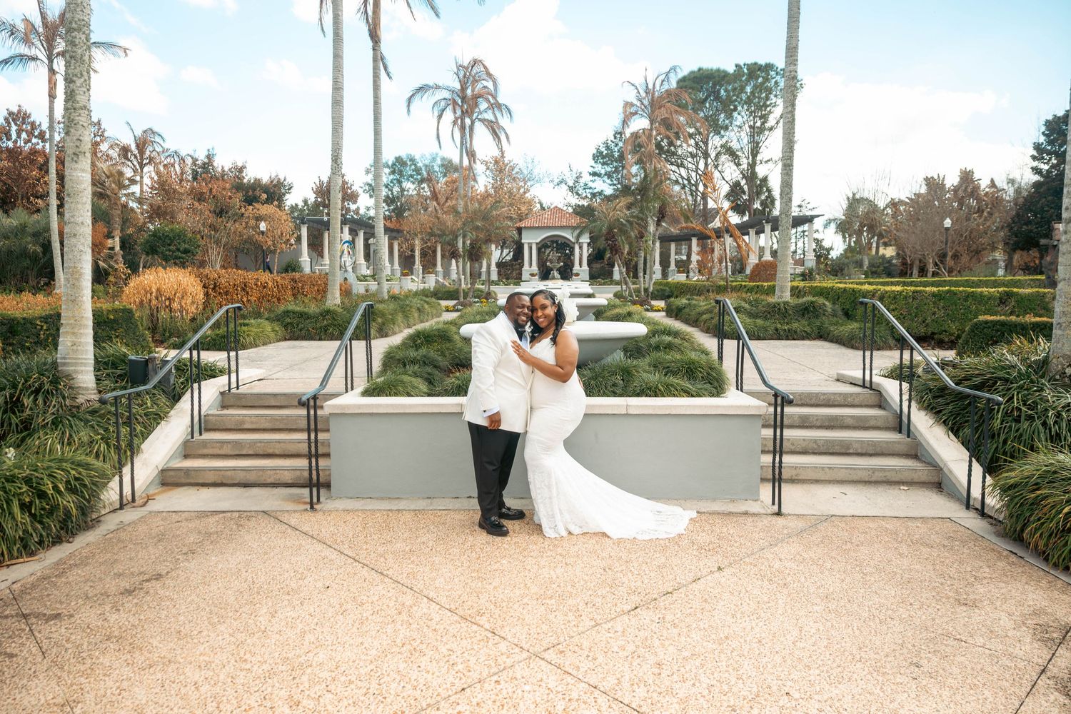 Bride and groom posing together on steps of an elegant garden venue with palm trees.