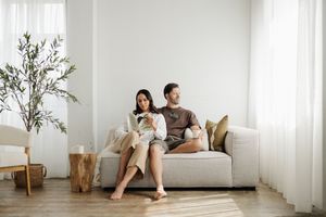 A couple relaxes on a white couch in a bright, minimalist living room with potted plants and natural decor.