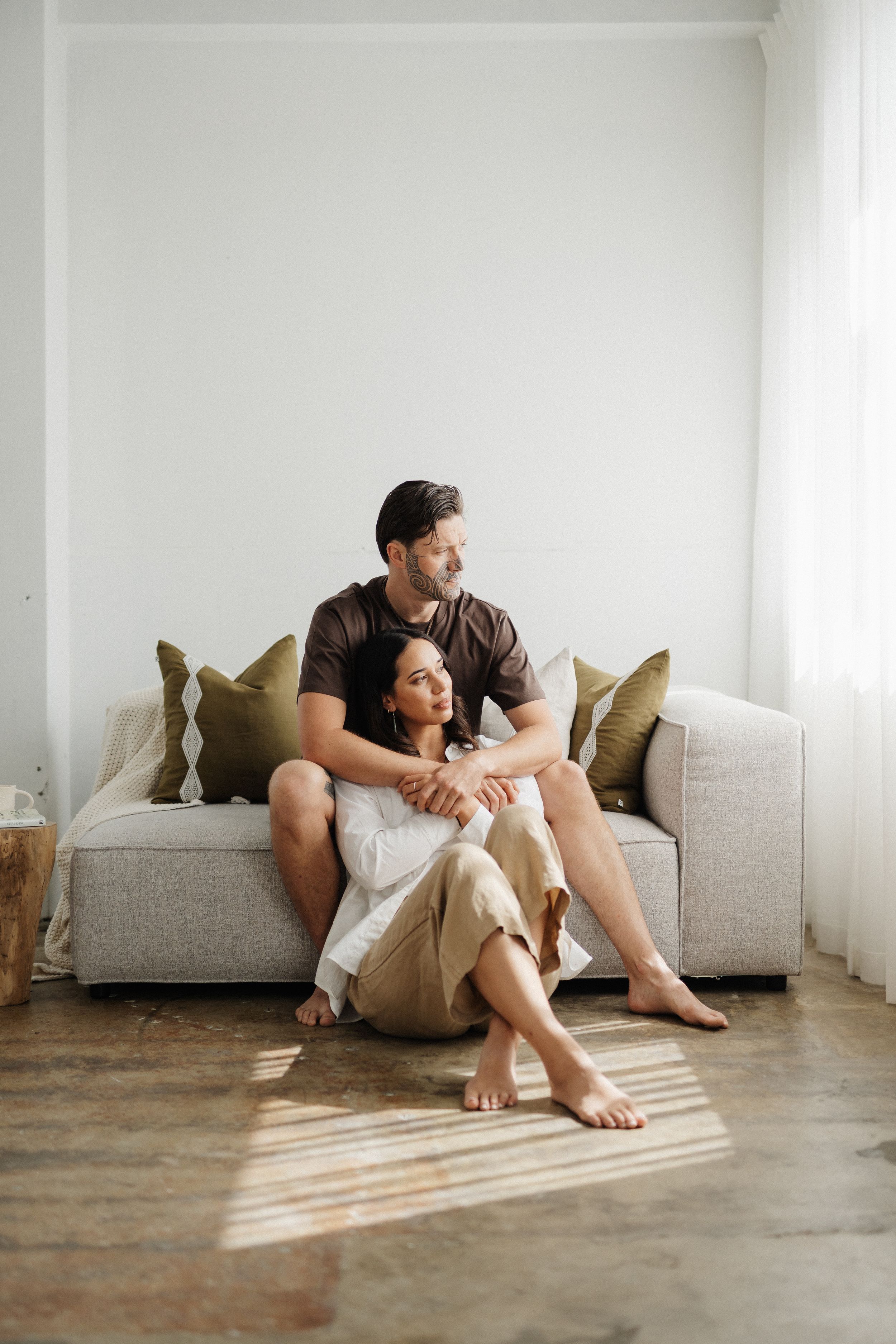 A couple sits comfortably together on the floor against a gray couch in a bright minimalist room with sunlight streaming in.