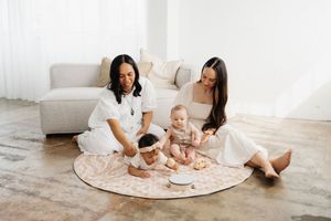 Two people sit on a white rug playing with a baby in a bright, minimalist living room setting.