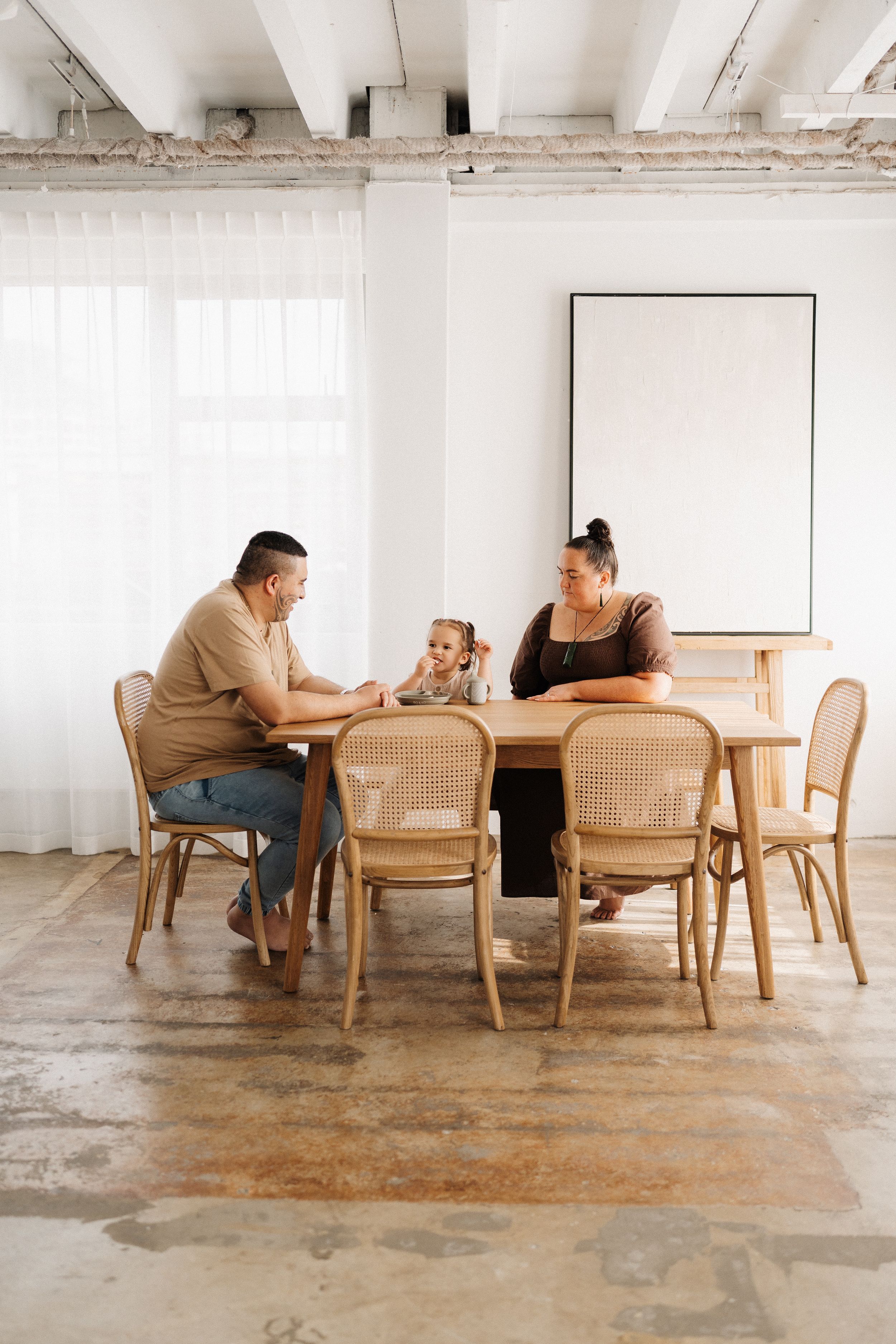 Family sitting together at wooden dining table with cane chairs in bright minimalist room with white walls and exposed beams.