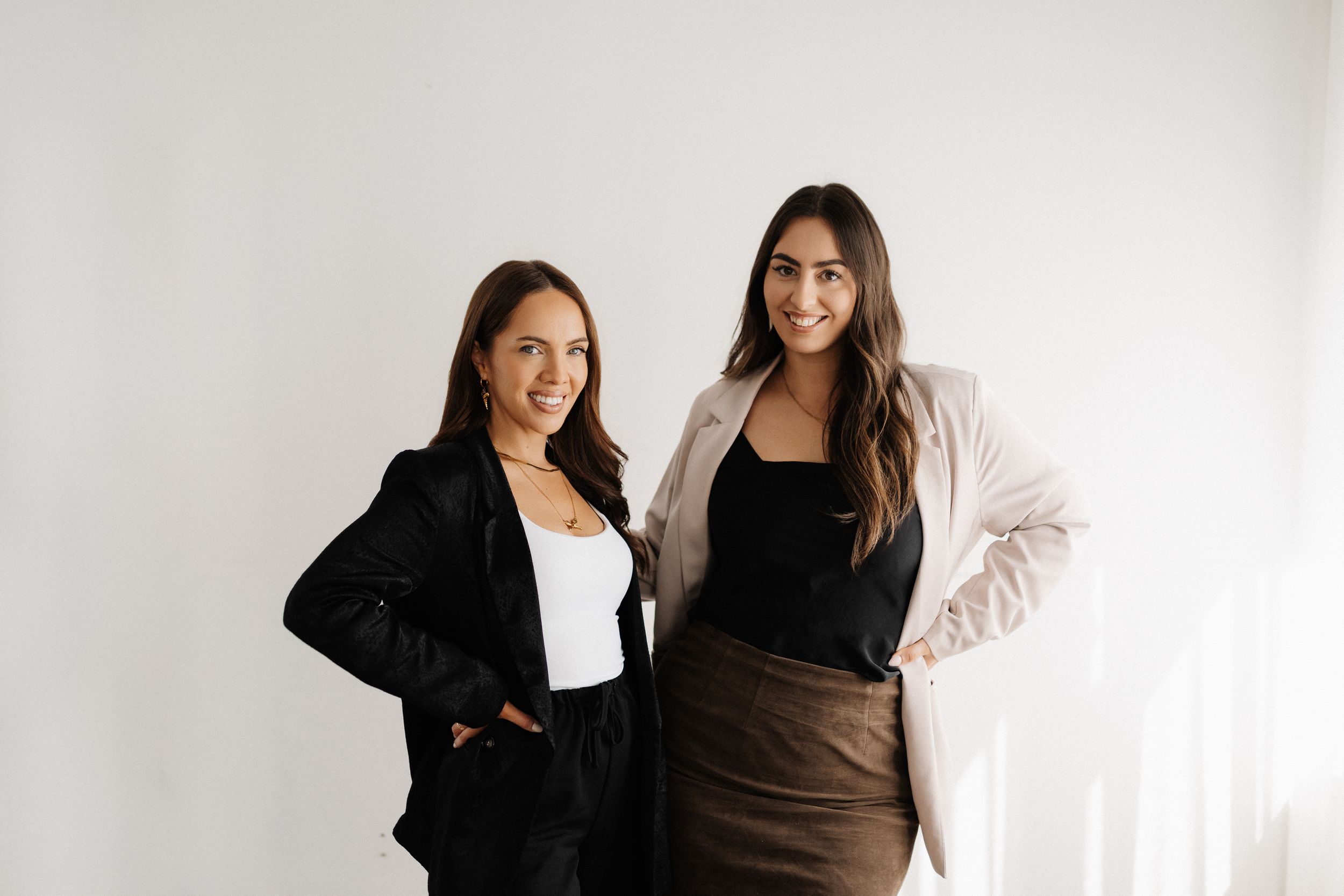 Two women in black and neutral toned outfits pose together against a white background.