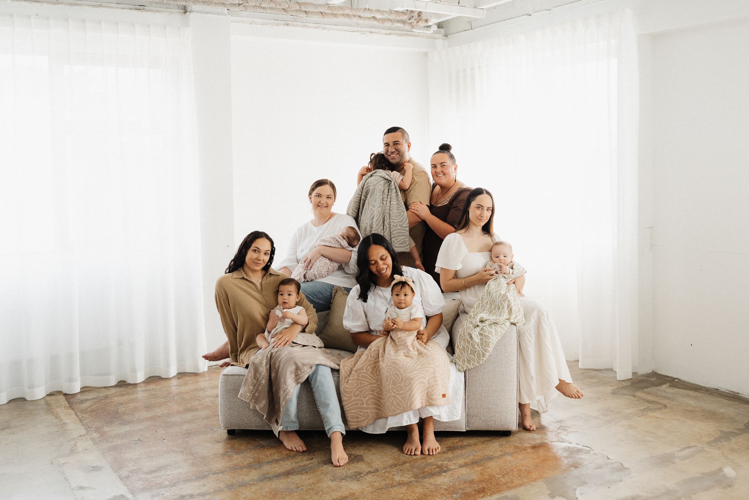 A group in coordinated neutral outfits poses together on a couch in a bright white studio setting.