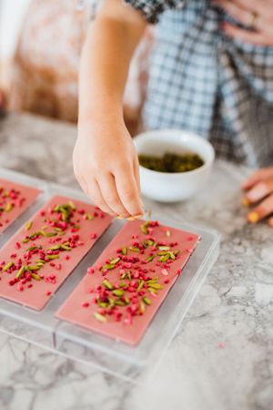 Pink chocolate bars being decorated with sprinkled pistachios on a marble surface.