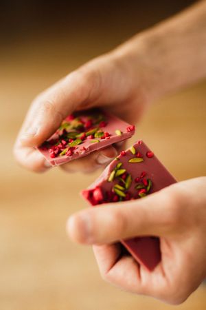 Hands holding pink chocolate with pistachio garnish against warm brown background.
