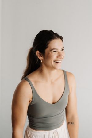 A person wearing a grey tank top poses against a white wall in a bright, airy setting.