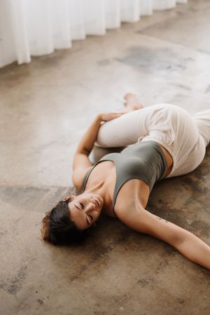 Person lying on concrete floor in workout attire doing reclined spinal twist yoga pose near white curtains.
