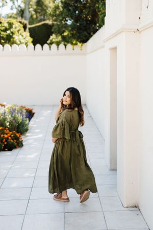 Woman in flowing olive green dress walking along white garden wall with blooming flowers.