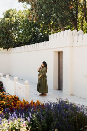 Person in long green dress standing by white wall with colorful garden flowers.