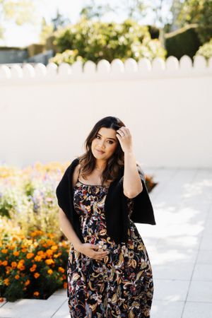 Woman in floral dress and black cardigan posing against white garden wall.