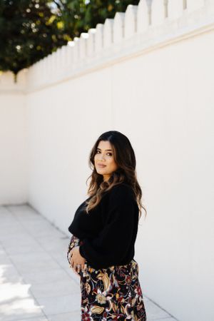 Portrait of woman in black cardigan and patterned dress against white wall background.