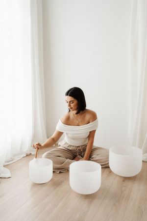 Person in off-shoulder outfit sits on floor with white crystal singing bowls.