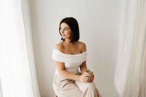 Person in white off-shoulder top sits on wooden stool against white backdrop.