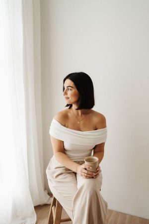 Person in white top holds beverage while seated on stool by window.
