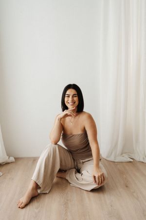 Person in neutral tones lounges on wooden floor against white curtain backdrop.