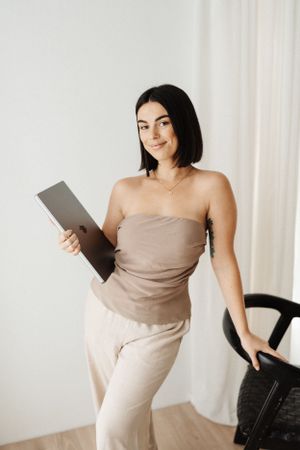Person in beige outfit holds tablet while standing in minimalist white room setting.