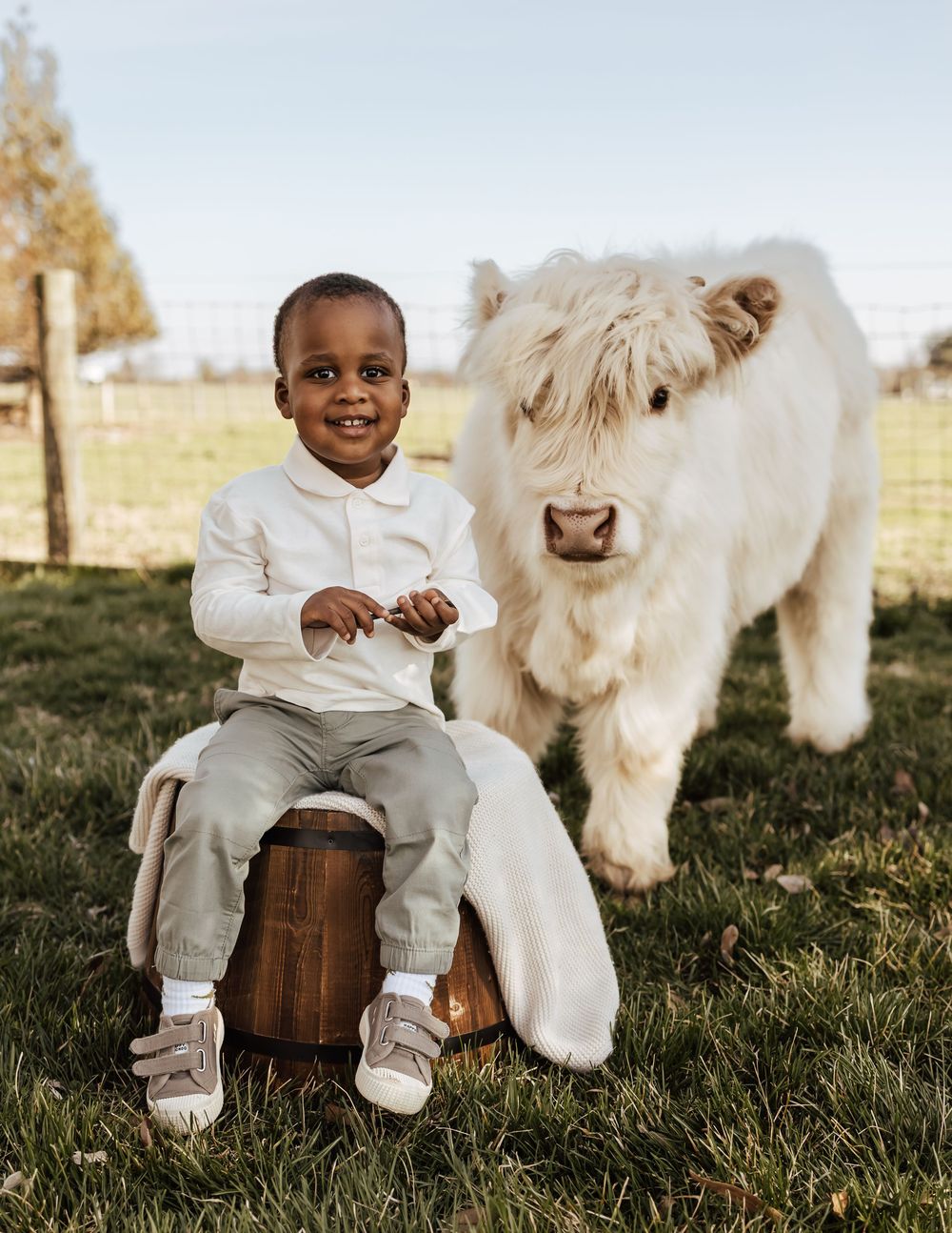 A child in white clothing sits on a wooden barrel beside a fluffy white dog in a grassy outdoor setting.